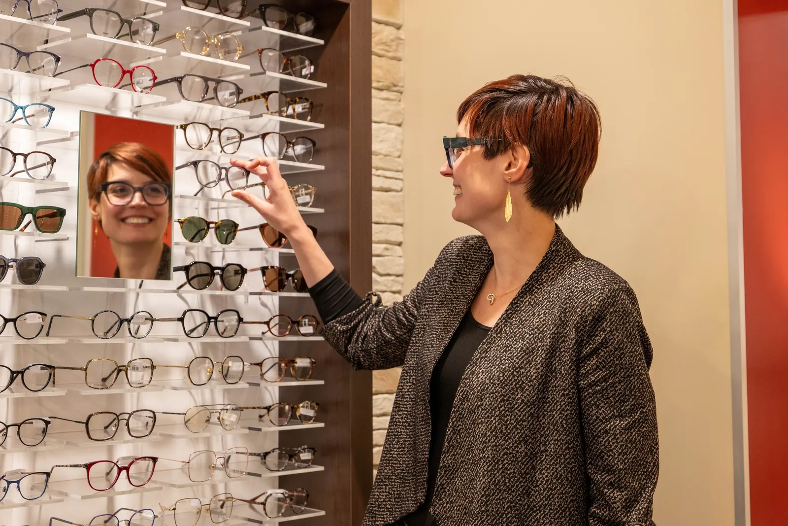 Opticienne conseillant une cliente devant le mur de montures de la boutique